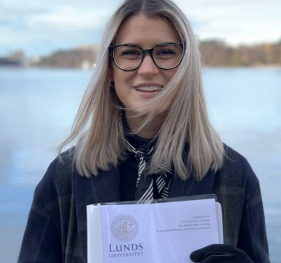 Josefine Svensson, som har axellångt blont hår och glasögon, håller upp sitt examensarbete. 